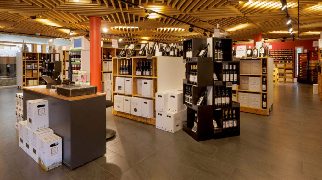 Weinladen Cave de La Côte mit Regalen voller Weinflaschen und Kartons in einem hell beleuchteten Raum mit roten Säulen