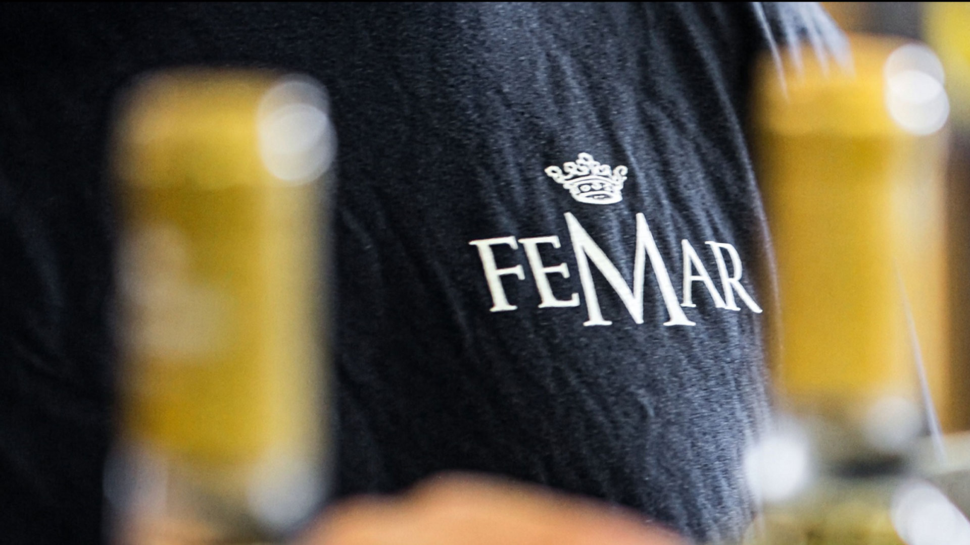 Schwarzes T-Shirt mit weissem Schriftzug 'FEMAR' und einer Krone darüber, unscharfe gelbe Flaschen im Vordergrund.