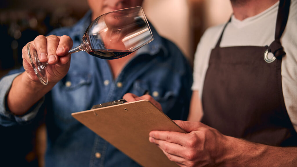 Zwei Männer begutachten die Weinqualität im Glas, während einer Notizen auf einem Klemmbrett macht.