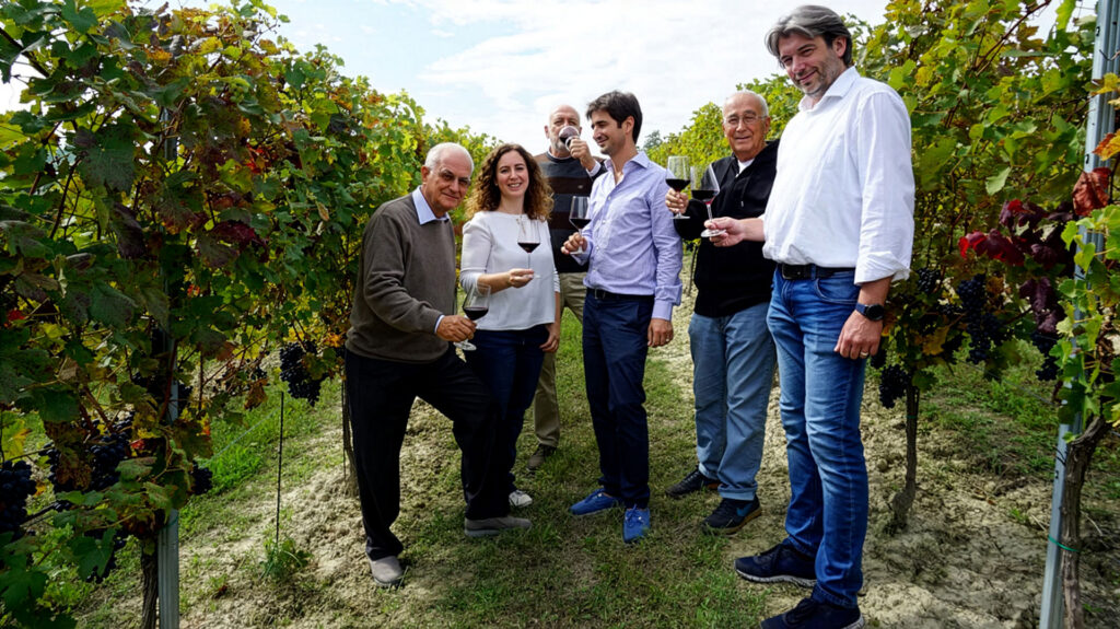 Fünf Personen stehen zwischen Weinreben und halten Weingläser in der Hand im Weingut Tenute Neirano