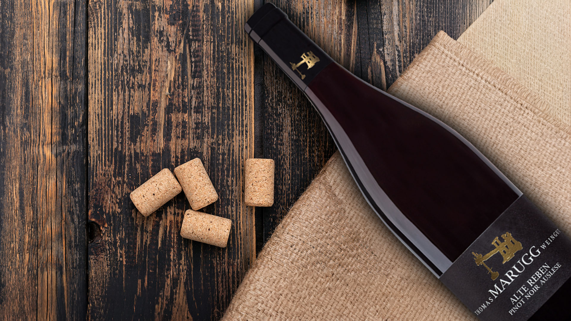 Flasche Wein mit schwarzem Etikett und goldener Schrift liegt auf grobem Leinenstoff neben mehreren Korken auf dunklem Holzuntergrund.