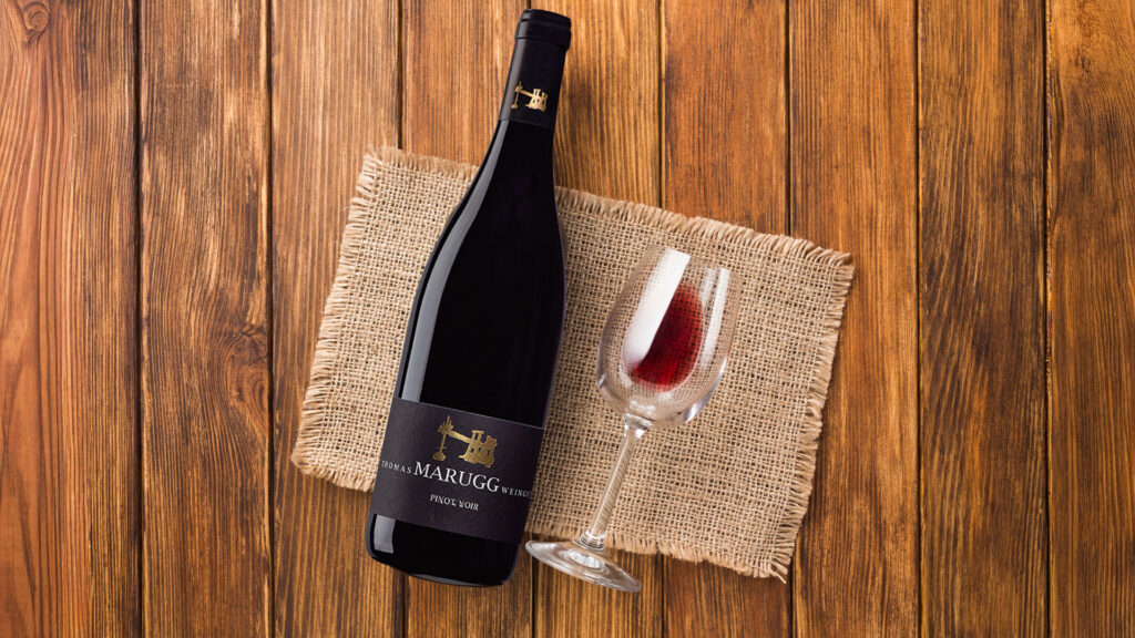 Flasche Pinot Noir von Weingut Thomas Marugg und halb gefülltes Weinglas auf grobem Leinentuch auf Holztisch.