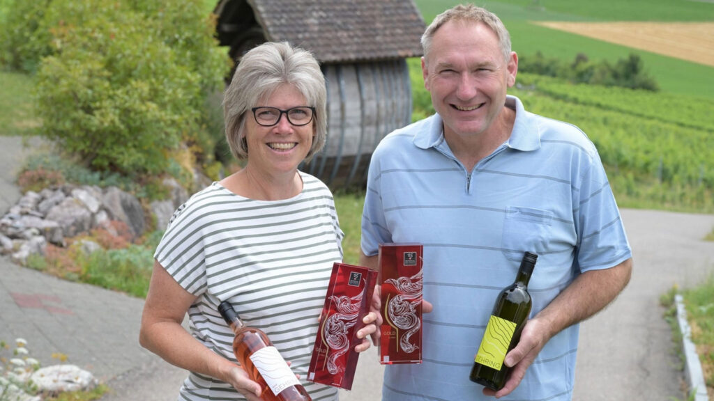 Peter und Yvonne Gehring steht im Freien, hält Weinflaschen und Geschenkverpackungen, im Hintergrund Weinberg und Holzhütte.