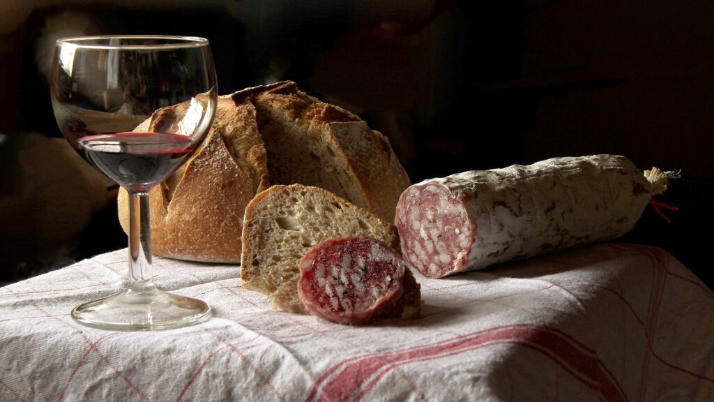 Weinglas mit Rotwein, rundes Brot mit zwei Scheiben und angeschnittener Salami auf weiss-rot kariertem Tuch.