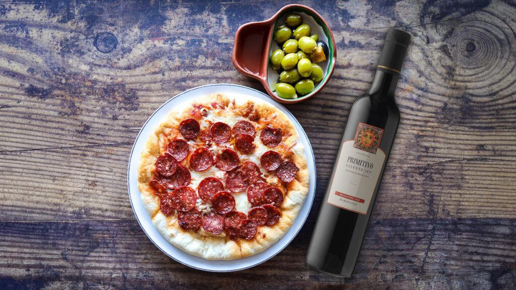 Pizza mit Salami auf weissem Teller, daneben Flasche Rotwein und Schale mit grünen Oliven auf Holztisch.