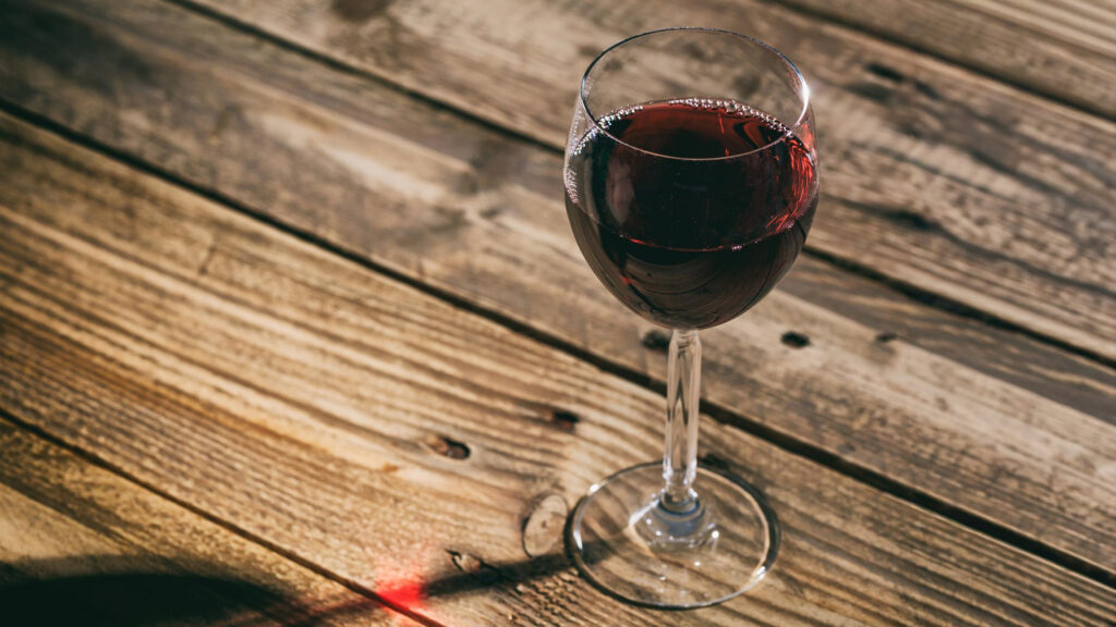 Weinglas mit rotem Wein auf einer Holzoberfläche, Lichtreflexe und Schatten sichtbar.
