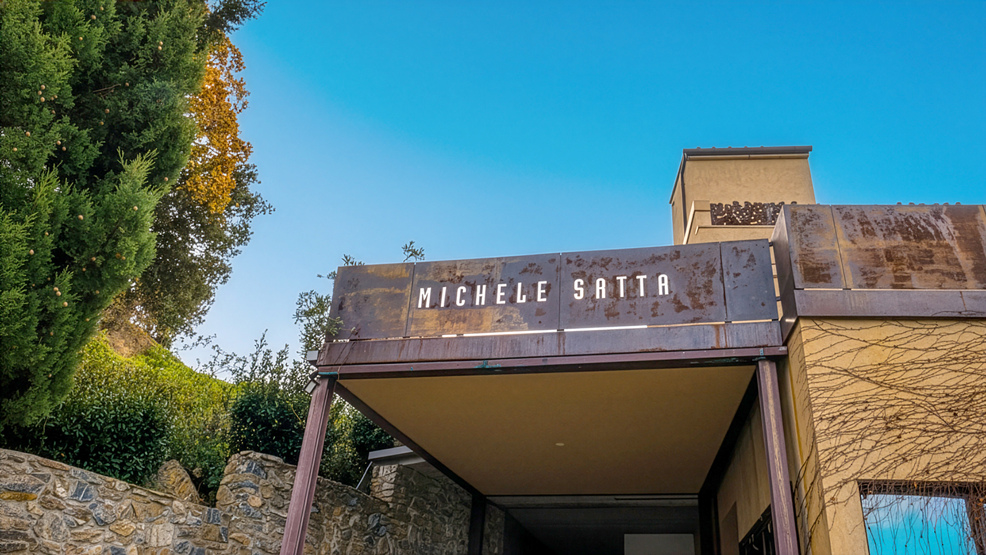 Eingang eines Gebäudes mit dem Schriftzug 'MICHELE SATTA' auf einer Metalltafel über dem Vordach, umgeben von Steinmauern und Bäumen.