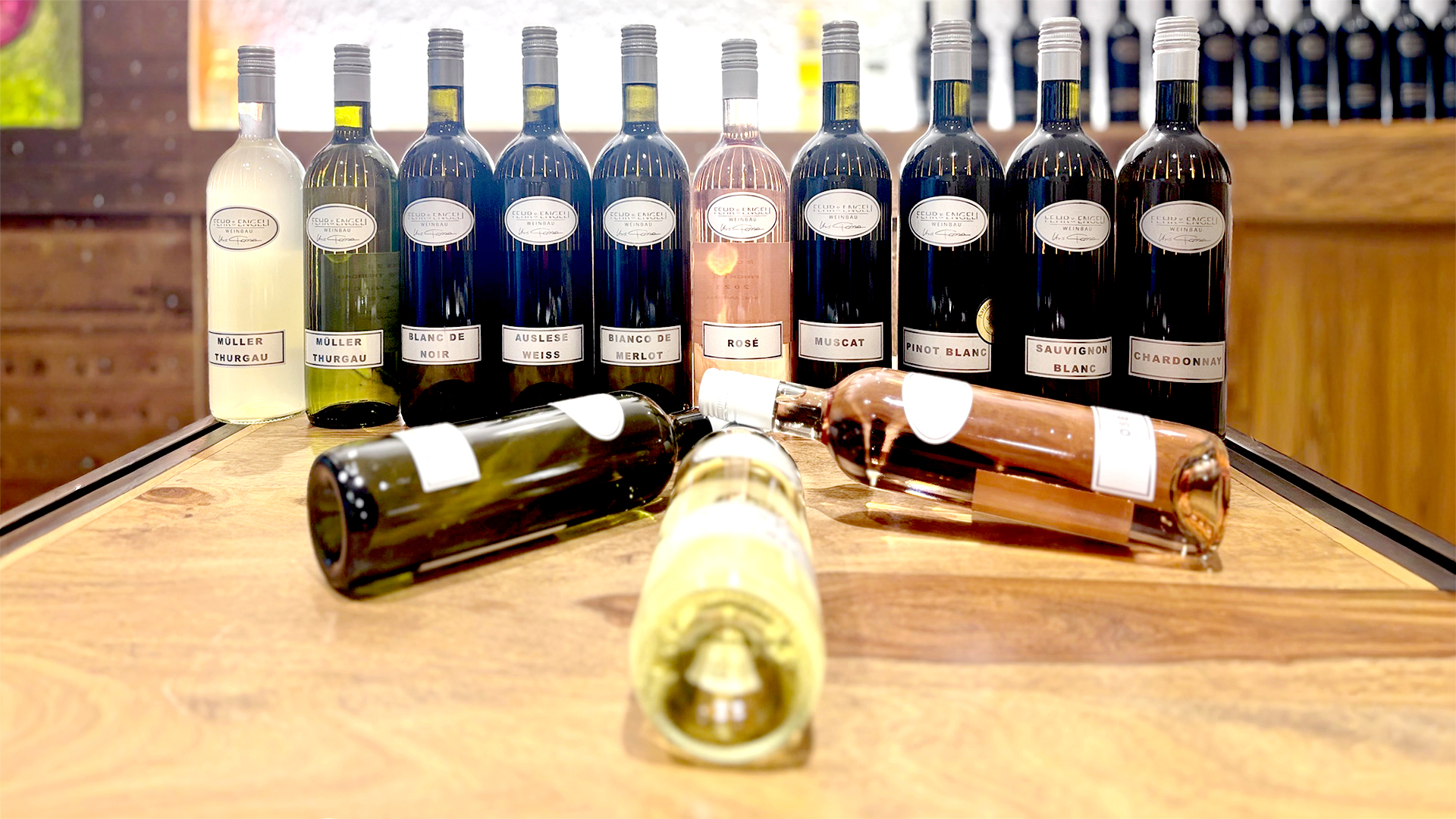 Mehrere Weinflaschen verschiedener Sorten stehen auf einem Holztisch, einige Flaschen liegen vorne flach, andere stehen im Hintergrund aufrecht.