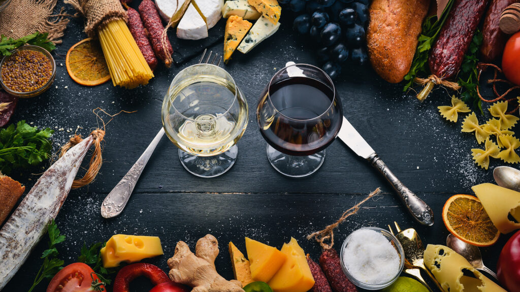Zwei Weingläser mit Weiss- und Rotwein auf dunklem Tisch, umgeben von Käse, Trauben, Brot, Pasta, Gewürzen und Wurstwaren.