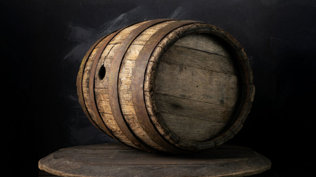 Altes Eichenfass mit Metallreifen liegt auf der Seite auf einem runden Holztisch vor dunklem Hintergrund.
