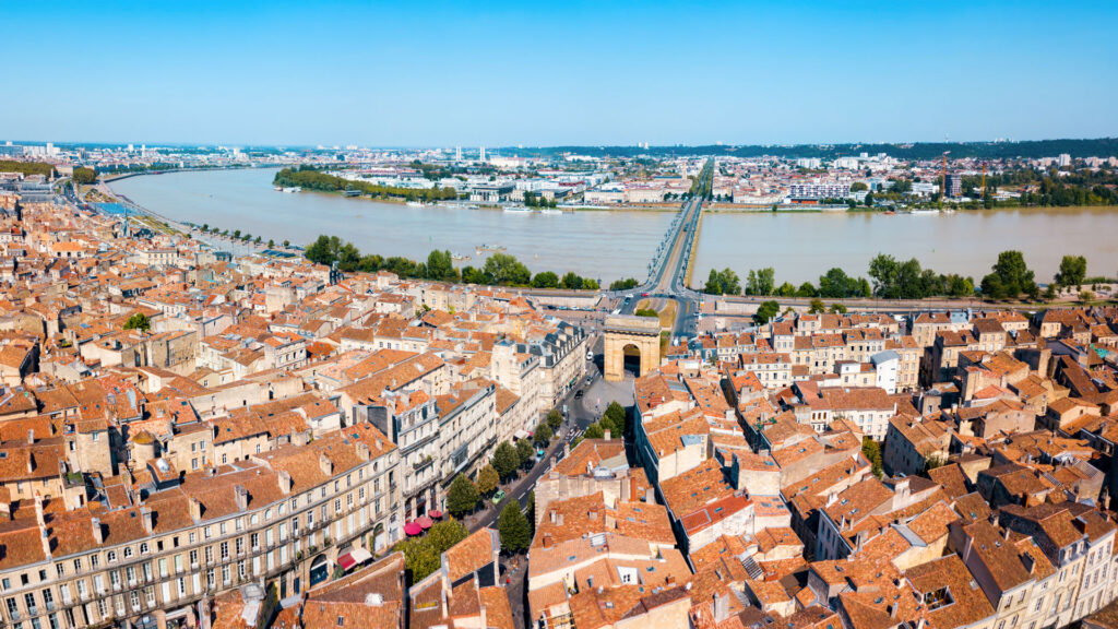 Luftaufnahme der Hafenstadt Bordeaux mit Fluss, Brücke und dicht bebauten Altstadtdächern mit roten Ziegeln.