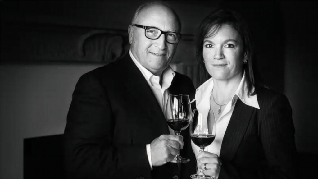 Zwei Personen in formeller Kleidung halten jeweils ein Weinglas mit Rotwein und stehen vor einem Kamin.