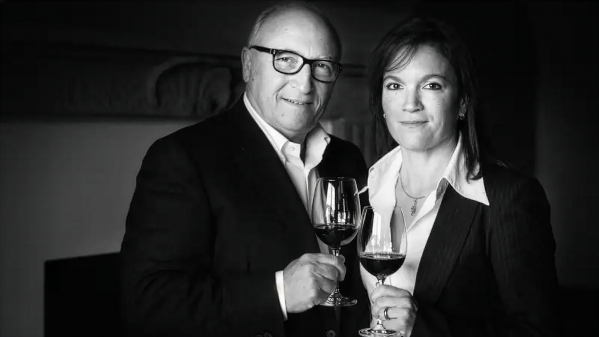 Zwei Personen in formeller Kleidung halten jeweils ein Weinglas mit Rotwein und stehen vor einem Kamin.