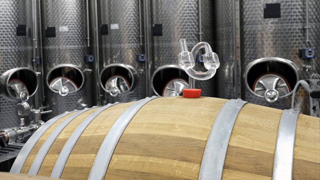 Holzfass mit Metallreifen und Gärspund im Vordergrund vor mehreren großen Edelstahlbehältern in einer Weinkellerei.