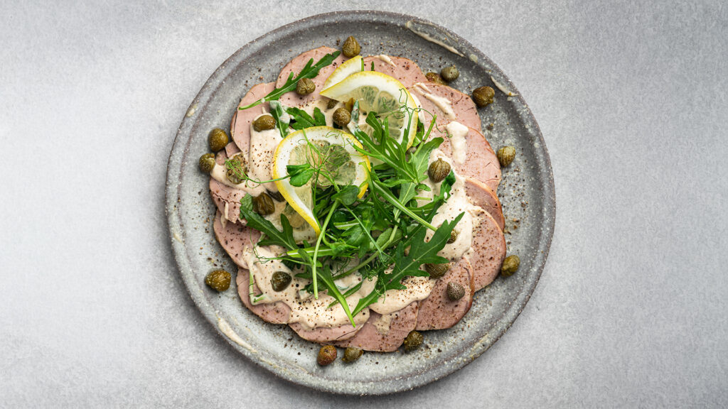 Rund angerichtete Scheiben von Vitello Tonnato mit Kapern, Zitronenscheiben und Rucola auf grauem Teller