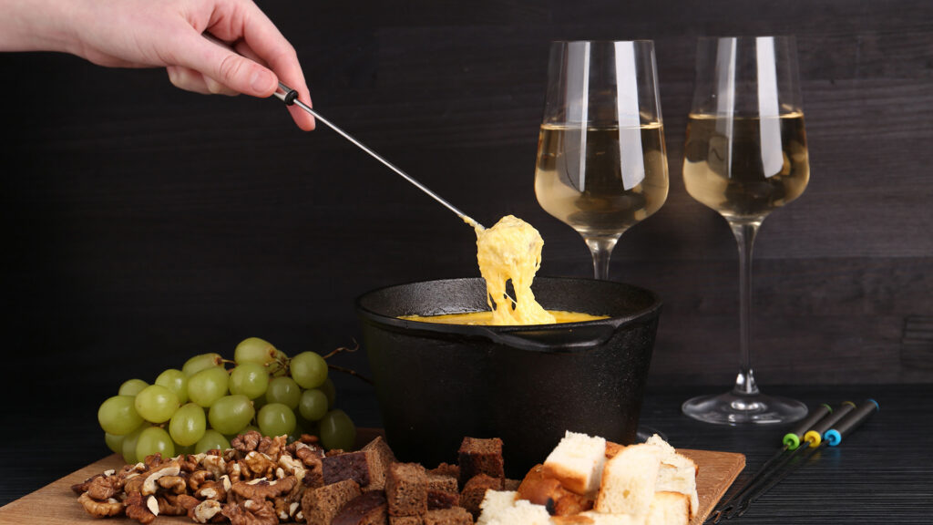 Auf einem Tisch steht ein schwarzer Caquelon gefüllt mit Käsefondue. Man erkennt oben links eine Hand, die mit der Fonduegabel ein Stück Brot umhüllt mit Käse aus dem Topf hebt. Rechts im Hintergrund stehen zwei Gläser gefüllt mit Weisswein. Vor dem Caquelon ist ein Holzbrett und darauf liegen verschiedene Brotsorten, Nüsse und weisse Trauben.