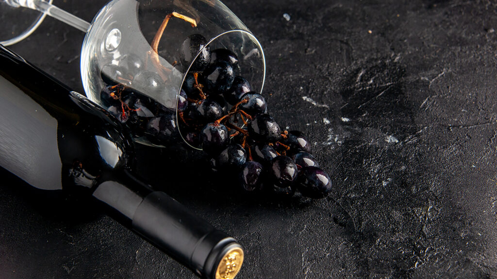Eine liegende Flasche Wein auf einem schwarzen Tisch, daneben ein liegendes Gas mit dunklen Trauben gefüllt