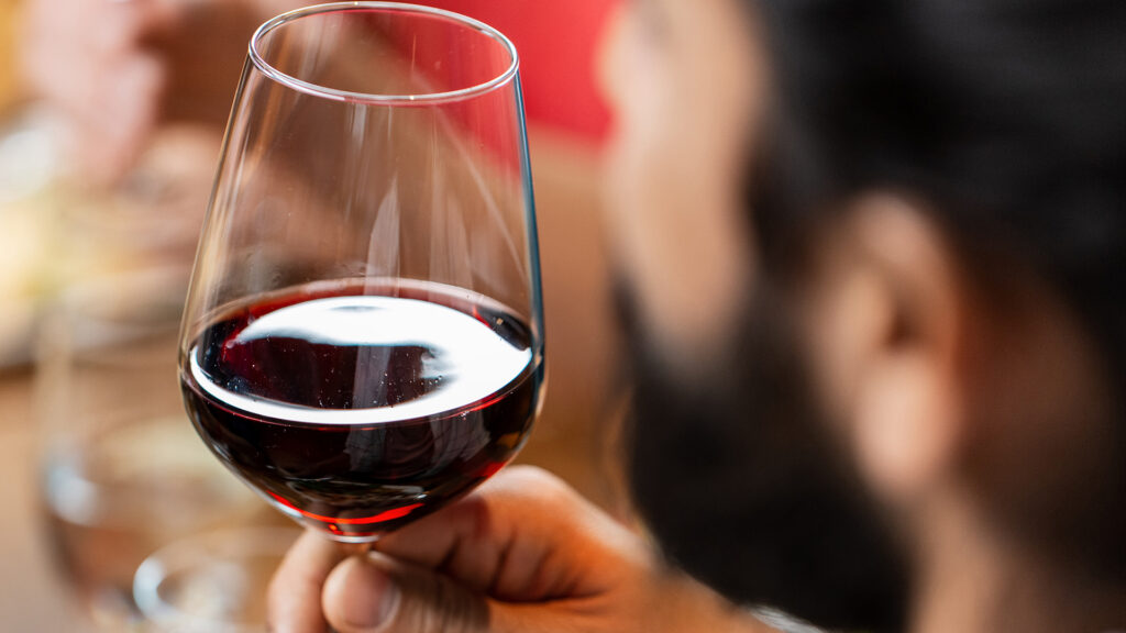 Ein Glas gefüllt mit Rotwein, das von einem Mann in der Hand gehalten wird. Man erkennt nur leicht das Profil seines Gesichtes, der Fokus ist auf dem Rotweinglas