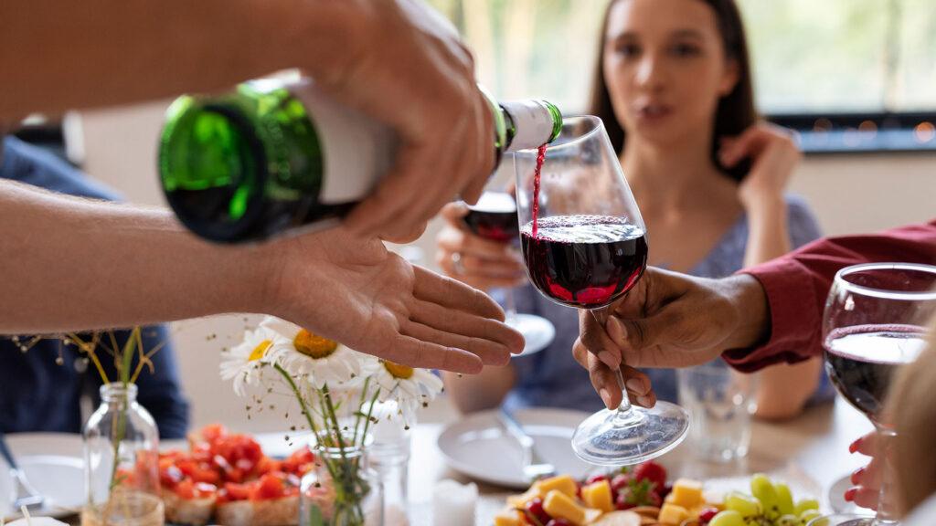 Ein Mann giesst ein Glas Rotwein ein. Am Tisch sitzen Gäste, auf dem Tisch steht Essen.